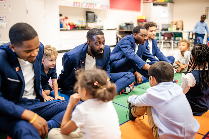 The MSU Denver Call Me MISTER students and director sit on a colorful mat in an elementary classroom across from young students, interacting and listening to what the students have to say.