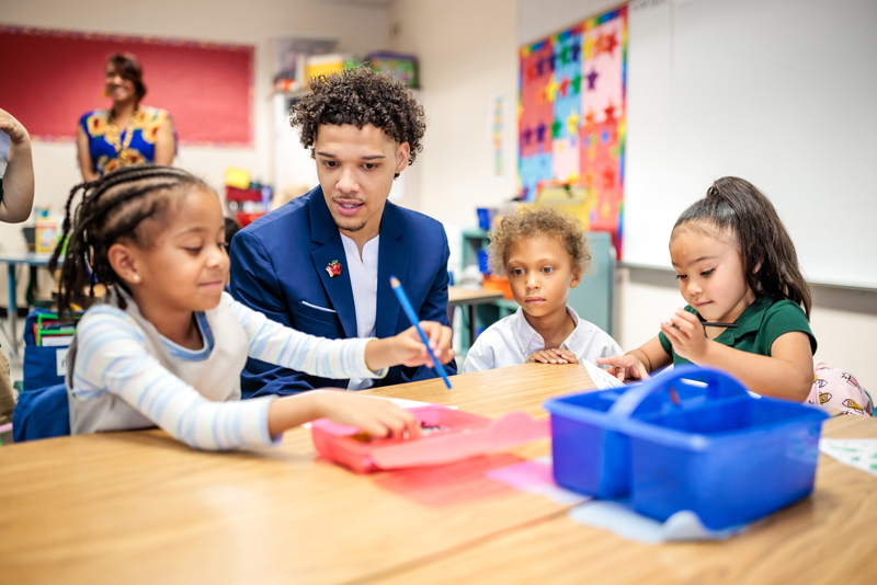 Jordan Puch, Call Me MISTER students, sits at a table in an elementary school classroom with three female students while they use colored pencils to draw.