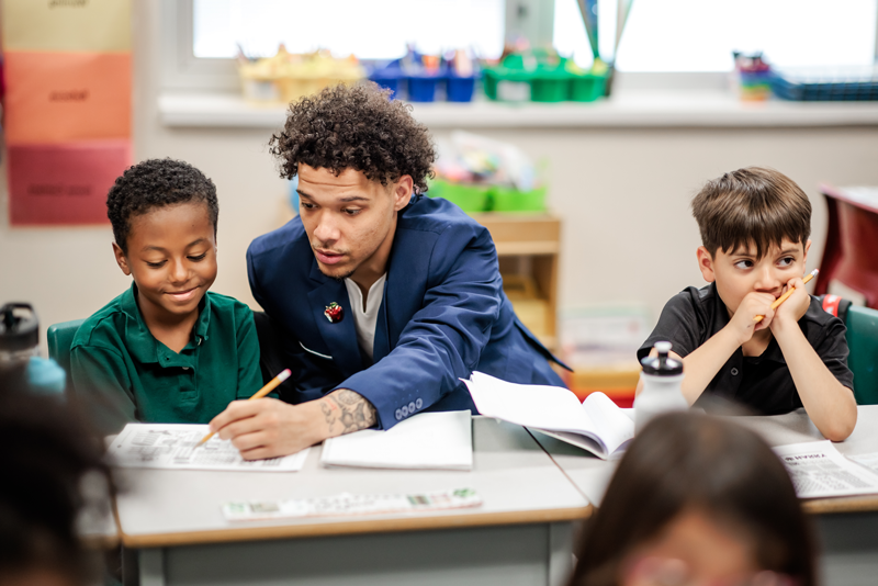 Jordan Puch, Call Me MISTER students, site at a desk in an elementary classroom between two young students, using a pencil to point to something in a workbook for one of the students while the student smiles.